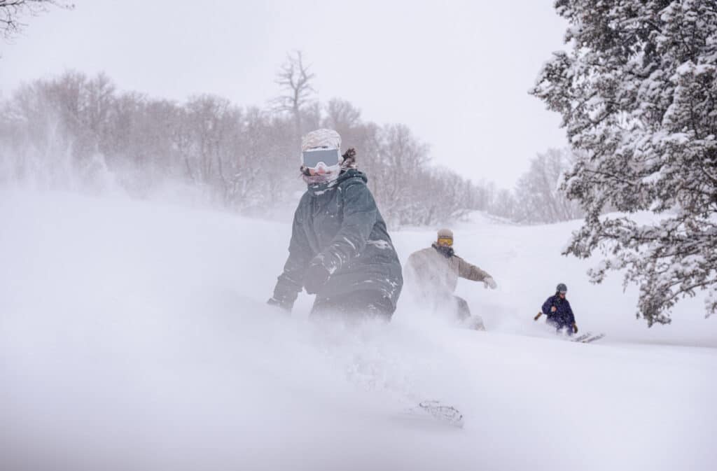 A group of skiers and snowboarders in deep snow. A rider in a black jacket leads the way